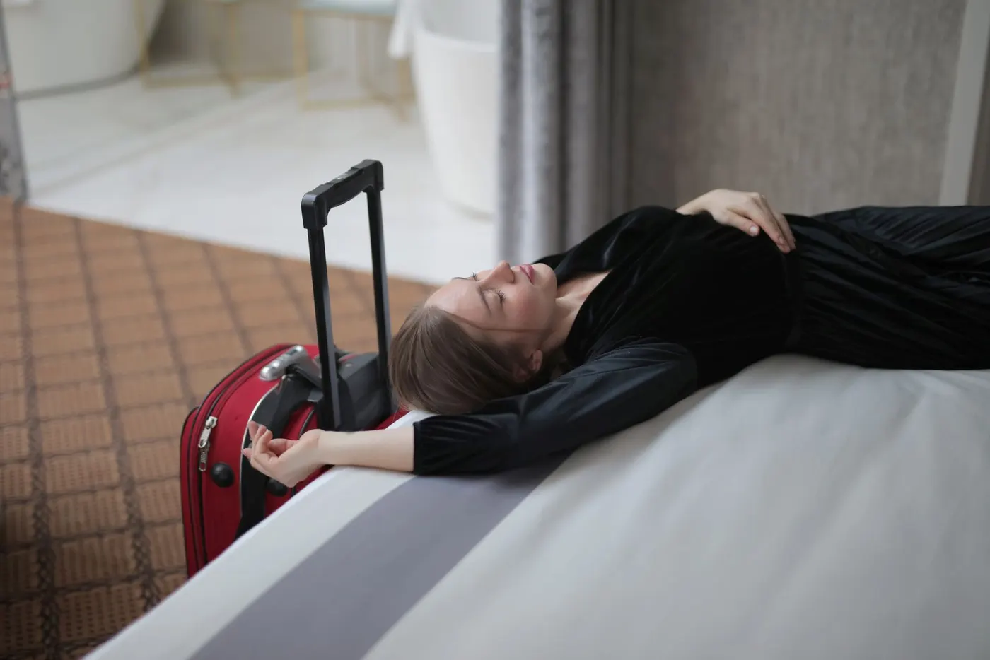 Una viajera agotada descansa en la cama de un hotel, junto a su maleta